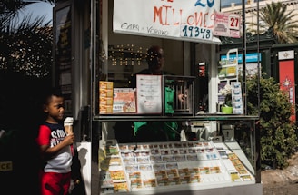 A child holding an ice cream cone stands outside a small kiosk or lottery ticket booth. The booth displays various colorful lottery tickets in the window. There are signs advertising a significant lottery prize amount in Spanish. A person can be seen partially obscured inside the booth, and plants are visible around the area, suggesting an outdoor setting.