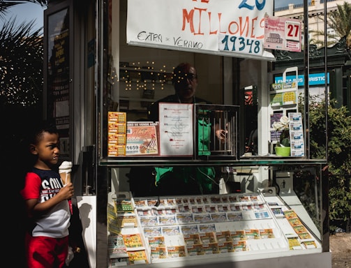 A child holding an ice cream cone stands outside a small kiosk or lottery ticket booth. The booth displays various colorful lottery tickets in the window. There are signs advertising a significant lottery prize amount in Spanish. A person can be seen partially obscured inside the booth, and plants are visible around the area, suggesting an outdoor setting.