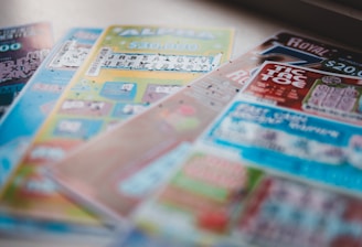 A selection of colorful lottery scratch-off tickets placed on a flat surface near a window. The image has a shallow depth of field, focusing on the tickets while blurring the background. The tickets display various games and prize amounts.