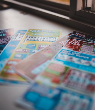 A selection of colorful lottery scratch-off tickets placed on a flat surface near a window. The image has a shallow depth of field, focusing on the tickets while blurring the background. The tickets display various games and prize amounts.