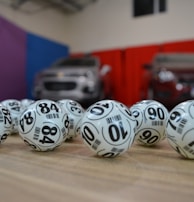 A collection of white lottery balls with black numerals scattered on a wooden surface. Two vehicles are blurred in the background, parked in a room with multicolored walls including teal, purple, blue, and red panels.